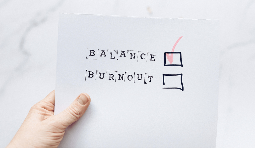 Eine Hand hält ein Papier mit den Wörtern Balance und Burnout. Balance ist angekreuzt.